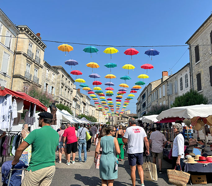 Marché estival de Nérac avec des parapluies colorés suspendus au-dessus des stands et des visiteurs.
